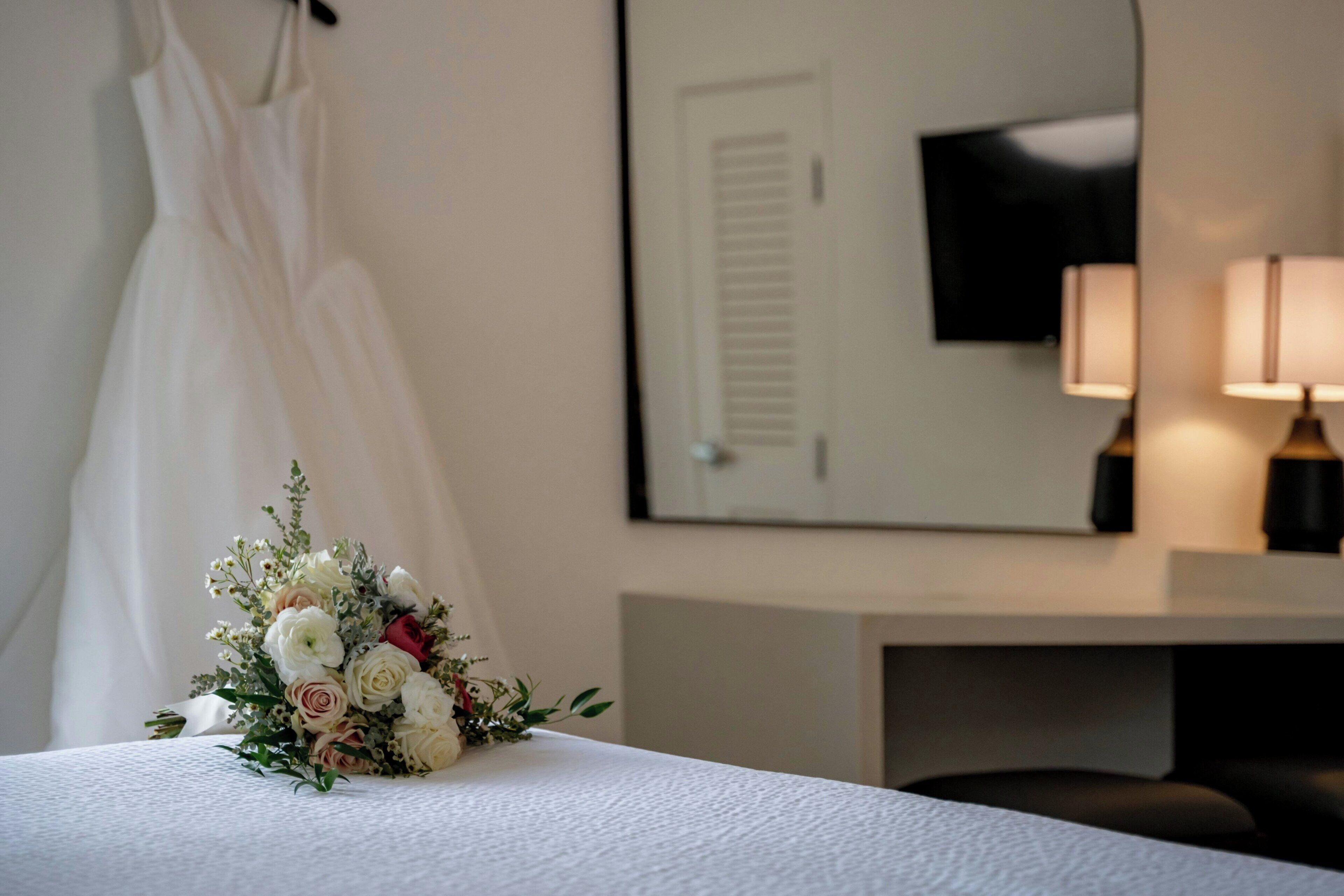 A hotel room with a neatly made bed, a bouquet of flowers on the bed, a hanging white curtain, a flat-screen TV mounted on the wall, and a bedside lamp.