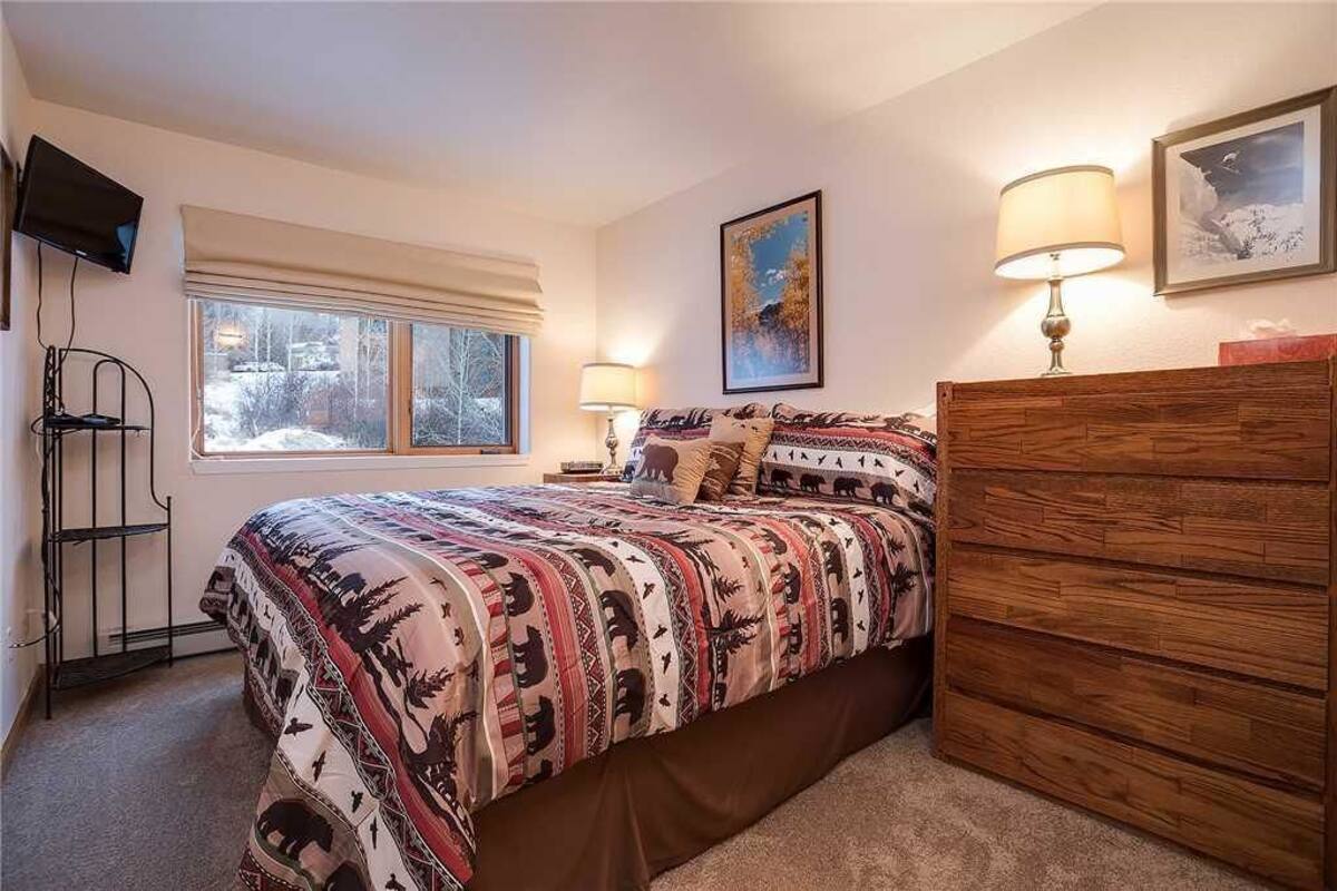 Une chambre à coucher avec un lit, des lampes de chevet, une commode, un téléviseur et une fenêtre donnant sur des arbres.