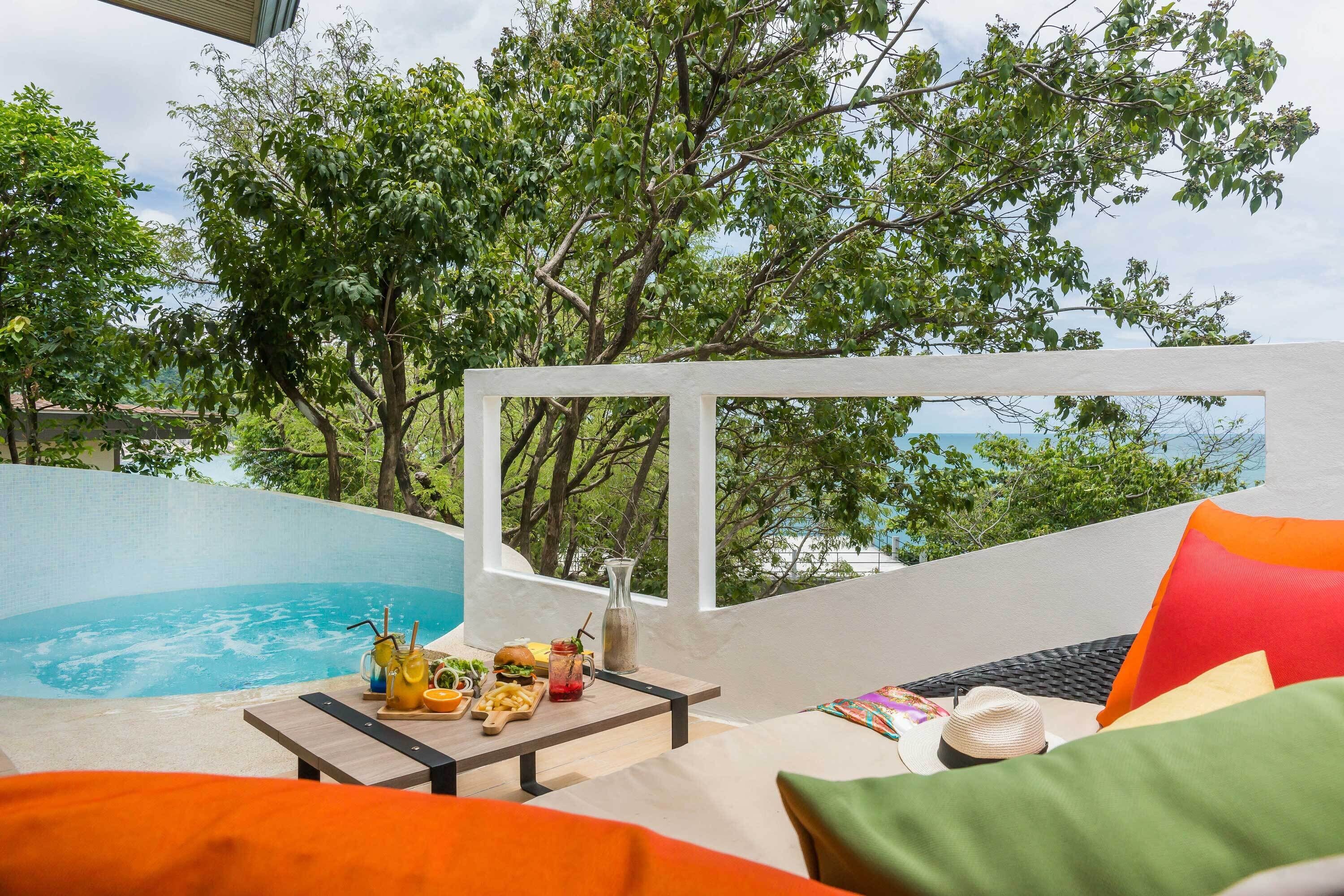 A poolside area with a table set for dining, a lounge chair, and a view of trees and a body of water.