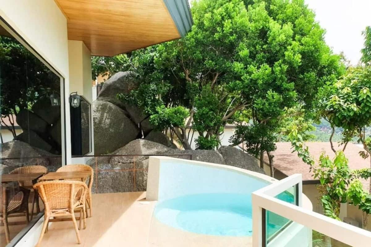 A pool area with a glass enclosure, wicker furniture, and a view of trees and rocks.