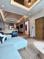 Una sala moderna con un sofá azul claro, una mesita de centro pequeña y un televisor montado en la pared. Se ve una puerta de madera y una zona de cocina al fondo.