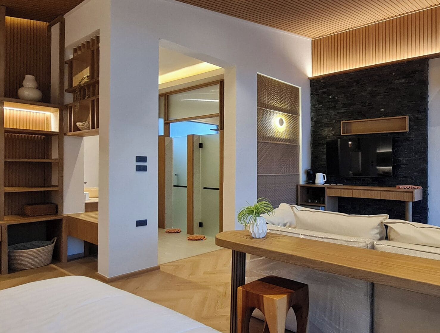 A modern hotel room with a wooden ceiling, a sofa, a dining table, and a TV mounted on a stone wall.