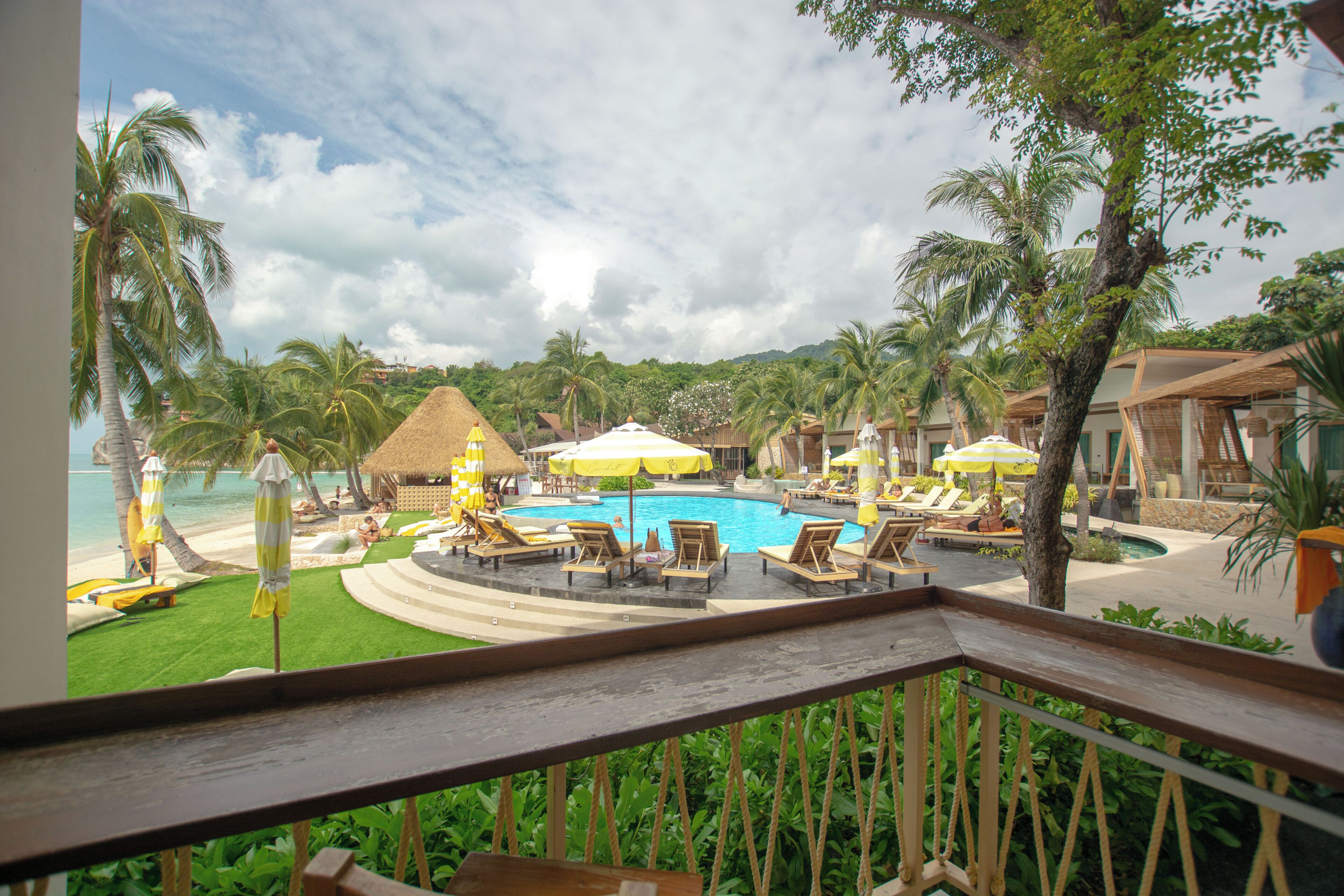 A resort pool area with lounge chairs and umbrellas, surrounded by palm trees and a view of the beach.