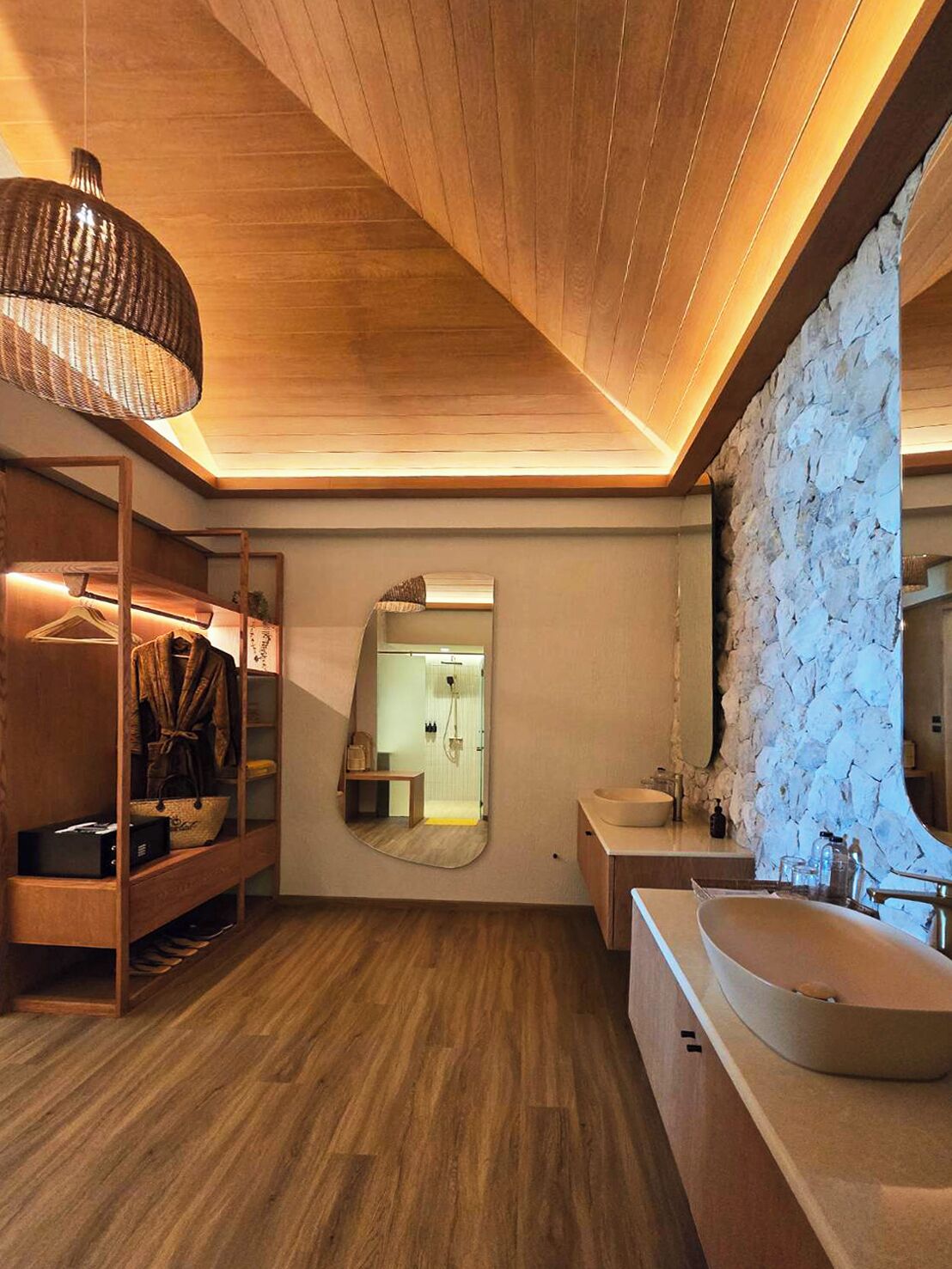 A modern bathroom with wooden flooring, a large mirror, a stone accent wall, and a wooden vanity with a sink.