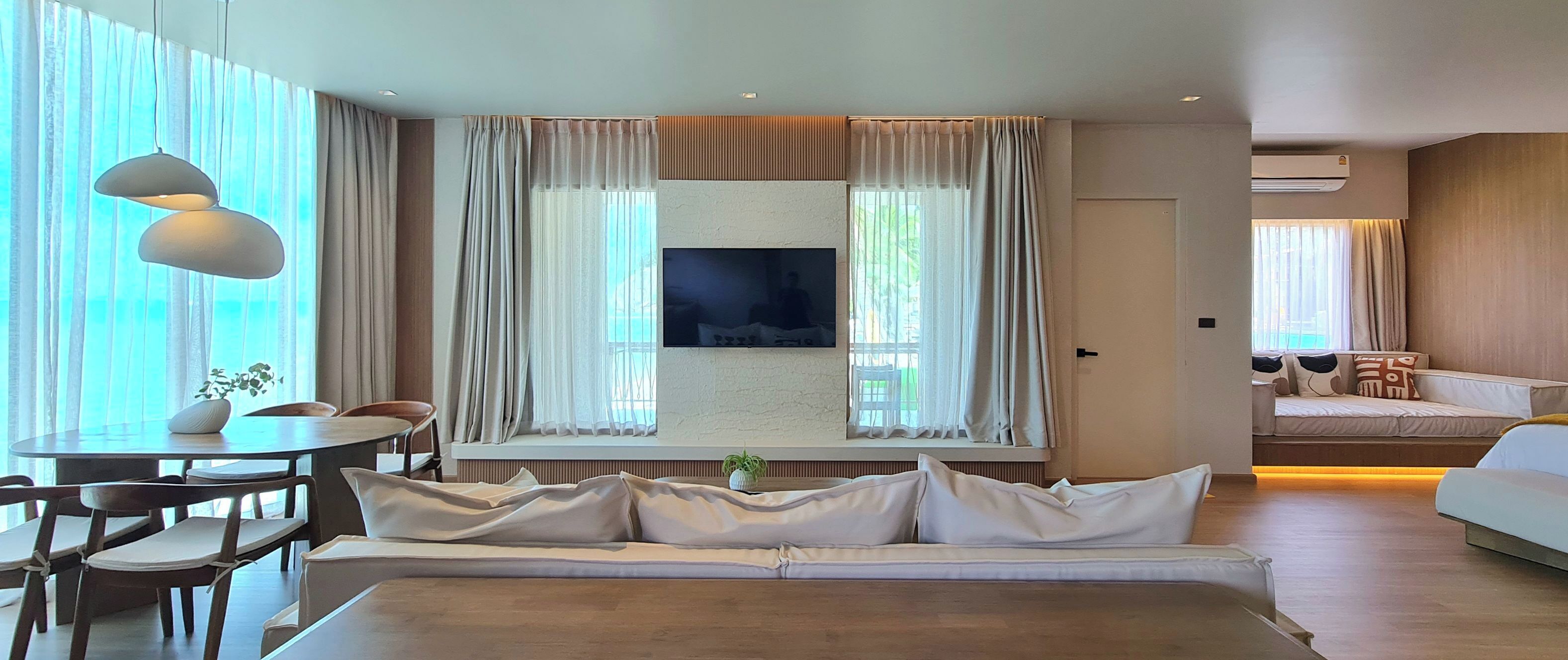 A modern hotel room with a large bed, a dining area with a table and chairs, a TV mounted on the wall, and a sofa in the corner.