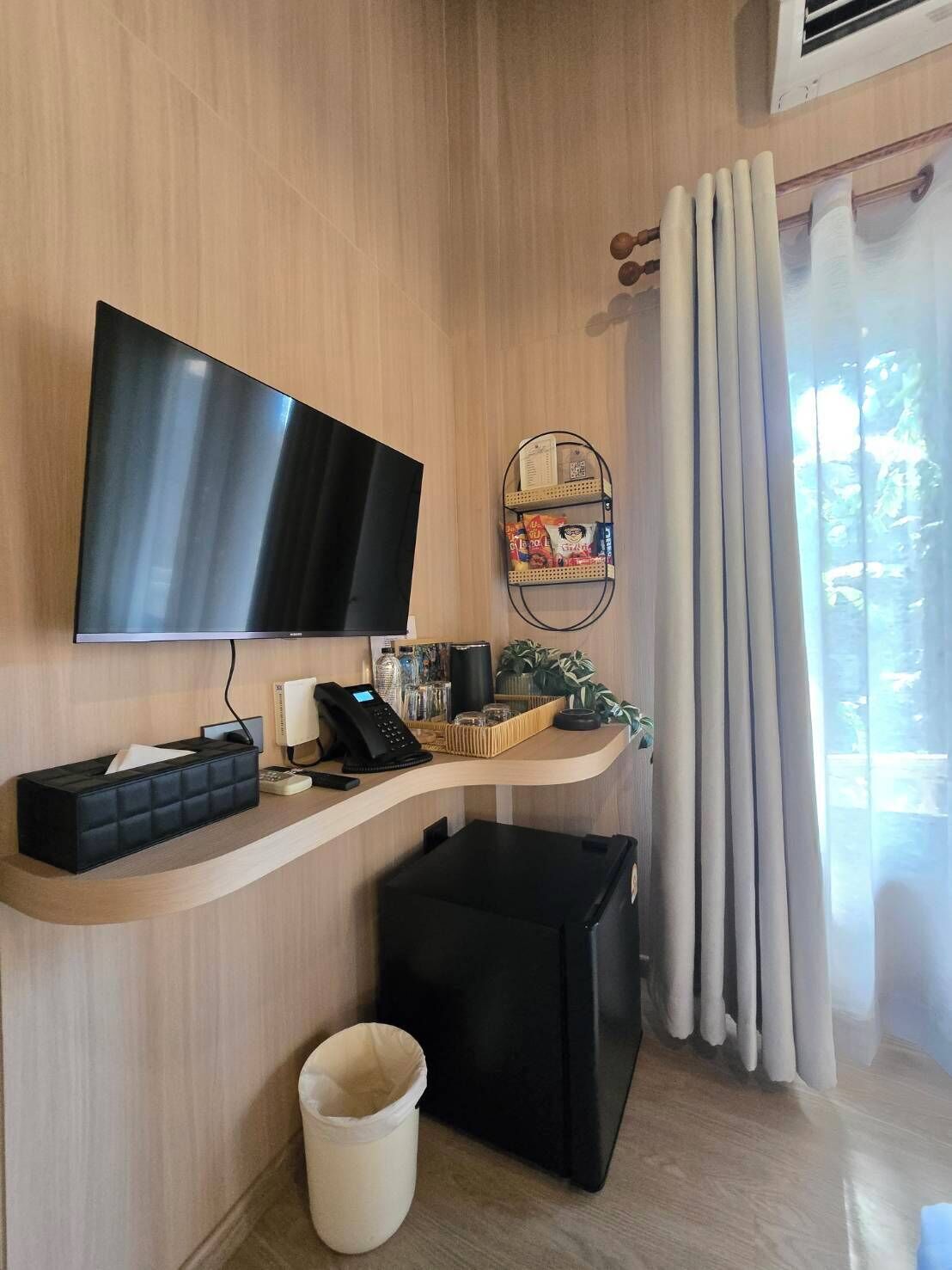 A modern room with a flat-screen TV, a small desk with a phone, a trash bin, and a window with curtains.