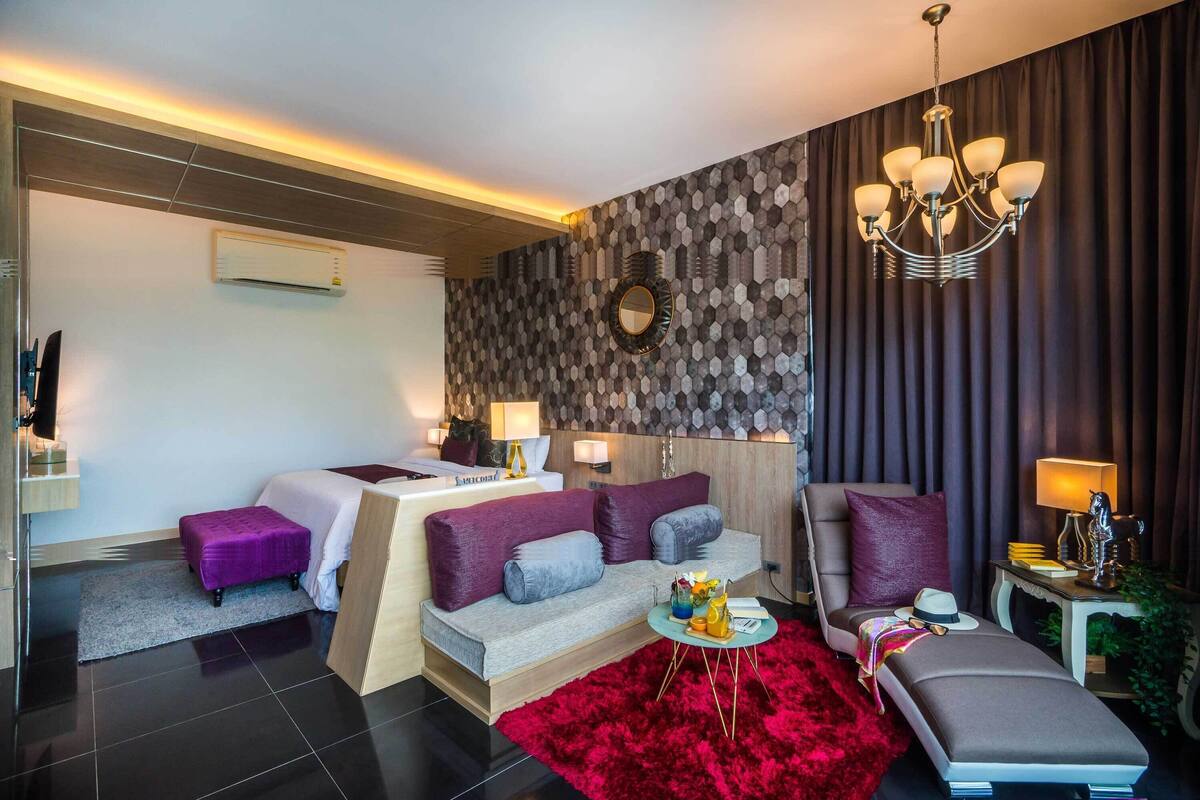 A modern hotel room with a sofa bed, a separate bed, a red rug, a chandelier, and a wall-mounted air conditioner.