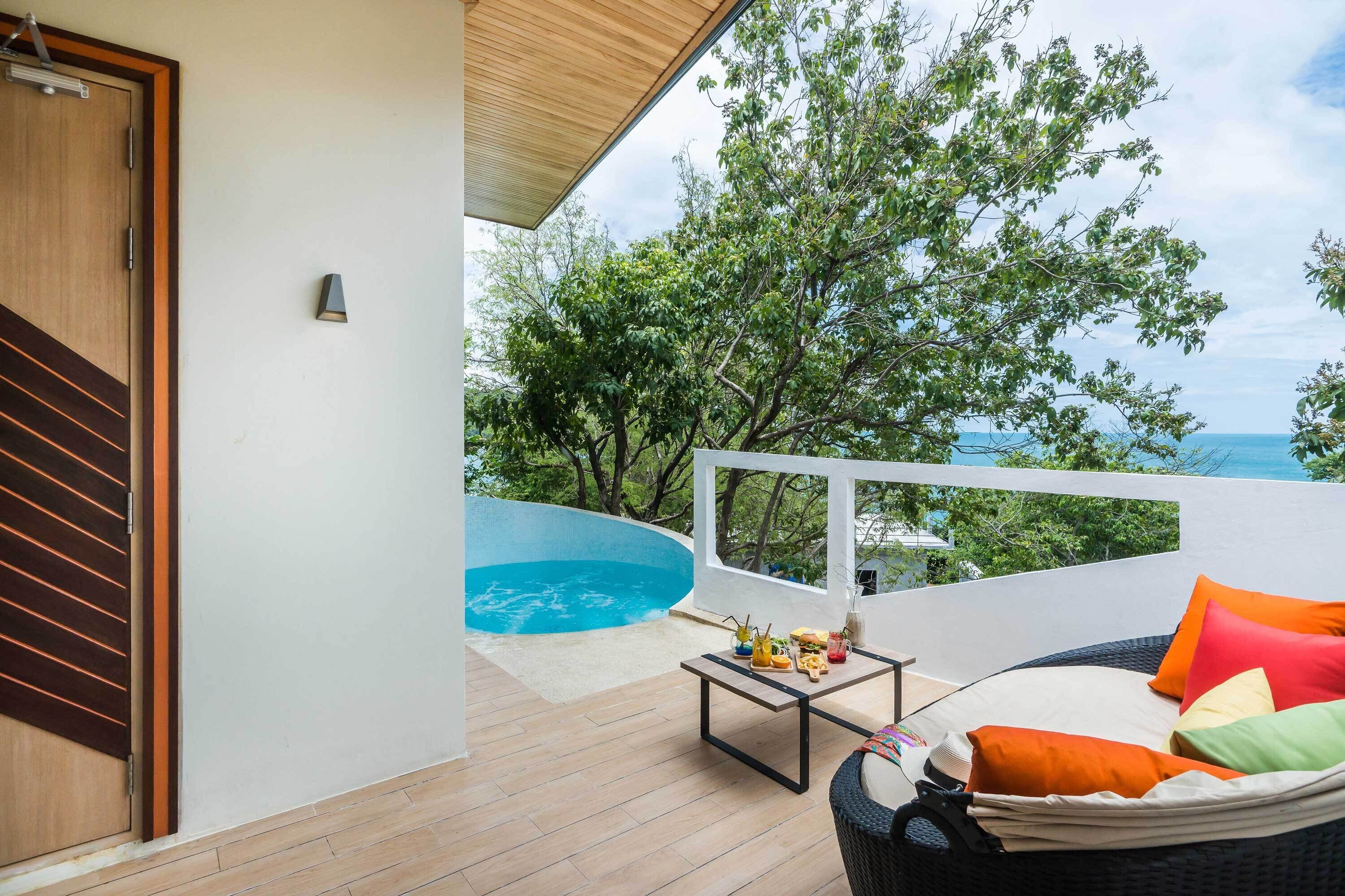 A balcony with a pool, wicker furniture, and a view of trees and the sea.