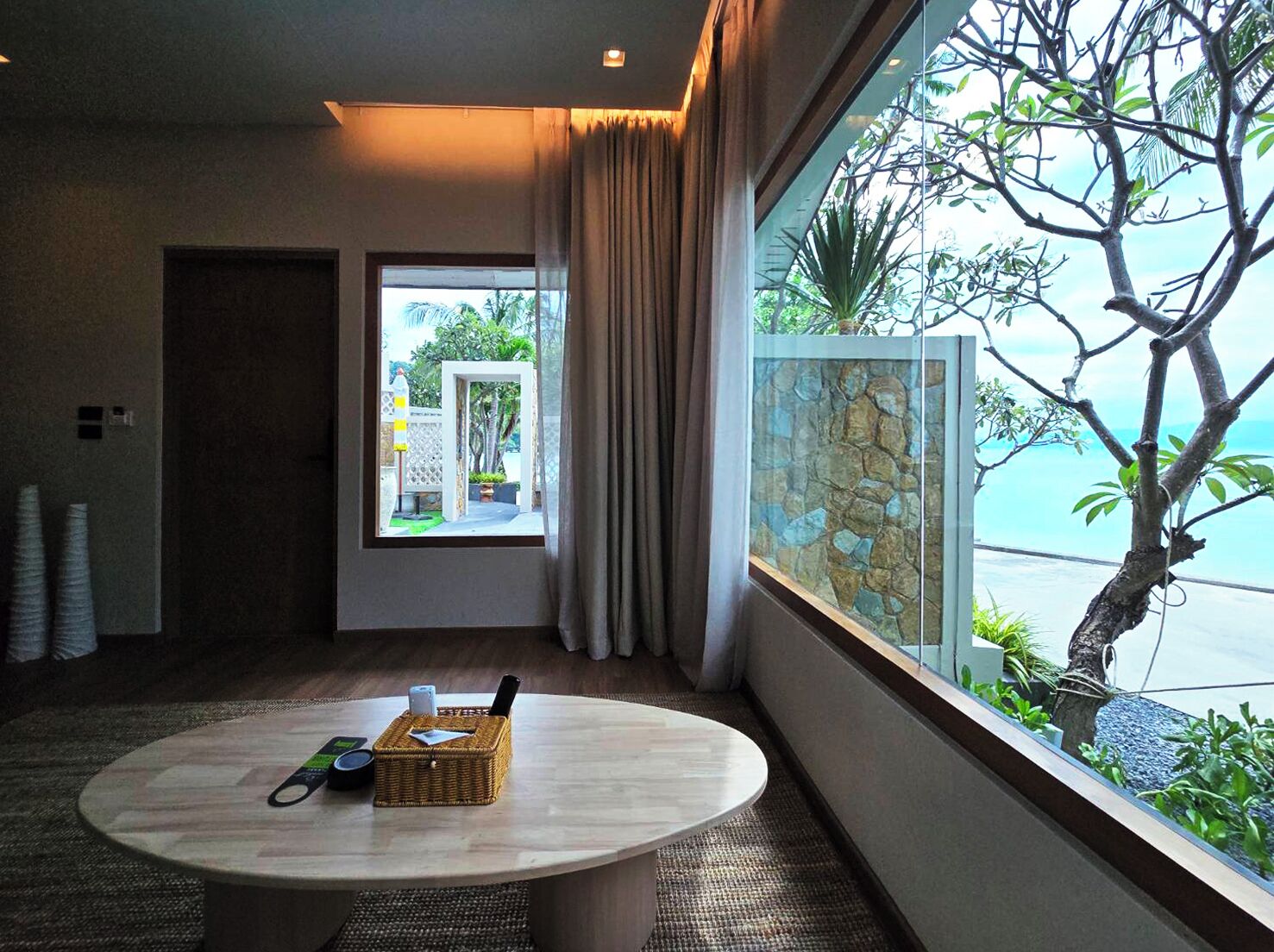 A hotel room with a round table, a wicker basket, and a view of a tree and a stone wall.