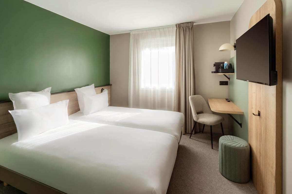 Quarto de hotel com duas camas, uma escrivaninha, uma cadeira, uma televisão e um pequeno banquinho.