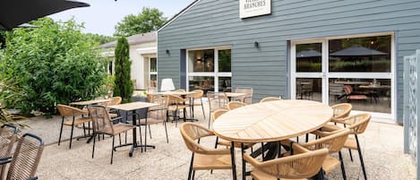 Un área de patio al aire libre con mobiliario de mimbre, un letrero que dice 'VIEILLES BRANCHES BUFFET BIEN FAIT', y un edificio con grandes ventanales.