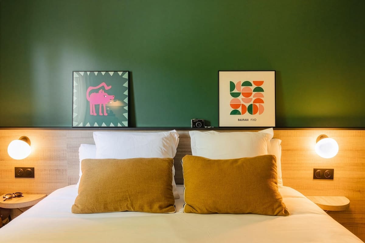 Quarto de hotel com cama, duas almofadas, mesa de cabeceira, uma câmera e duas obras de arte emolduradas na parede.