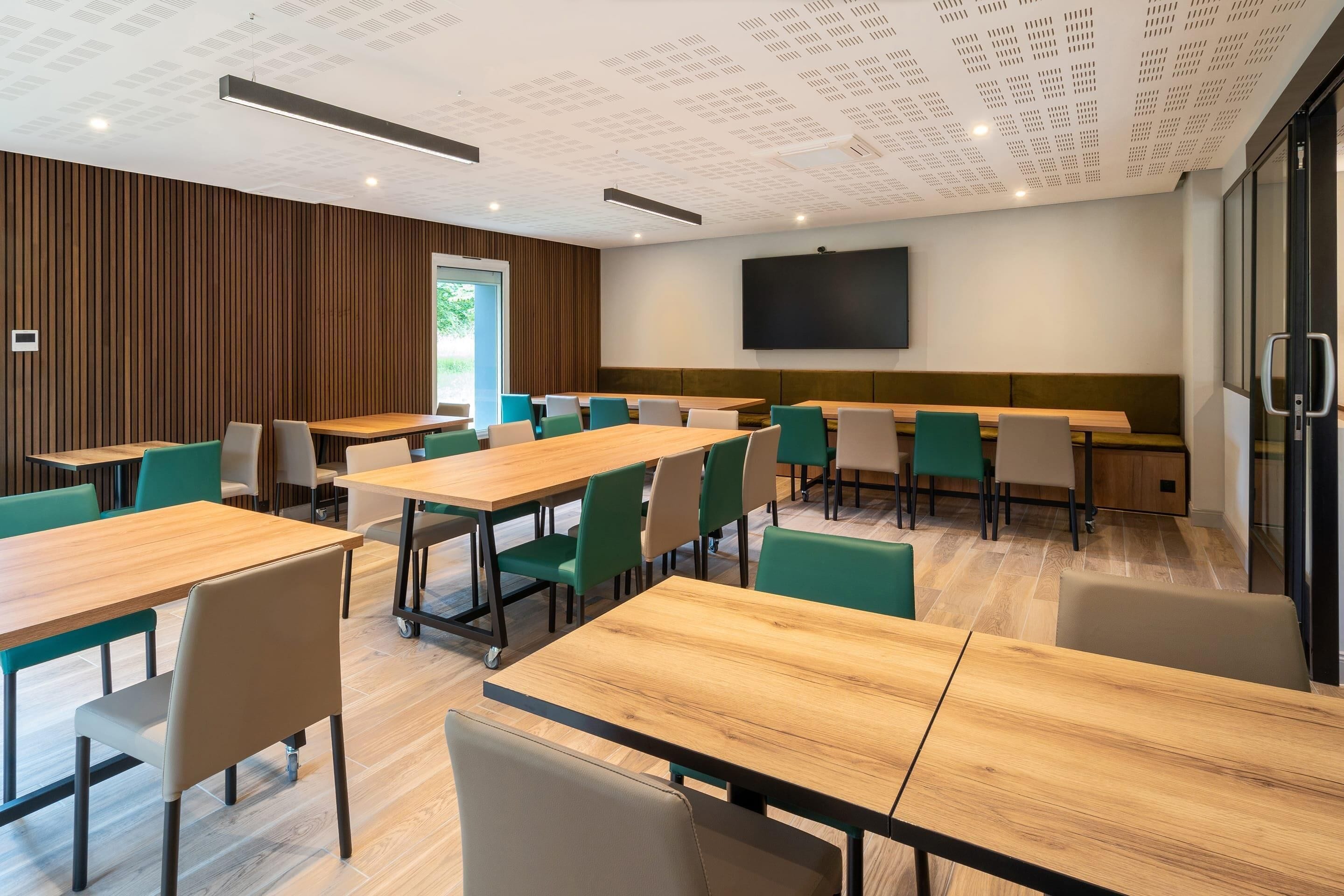 A modern conference room with wooden tables and green chairs, a large screen on the wall, and a wooden panel wall.