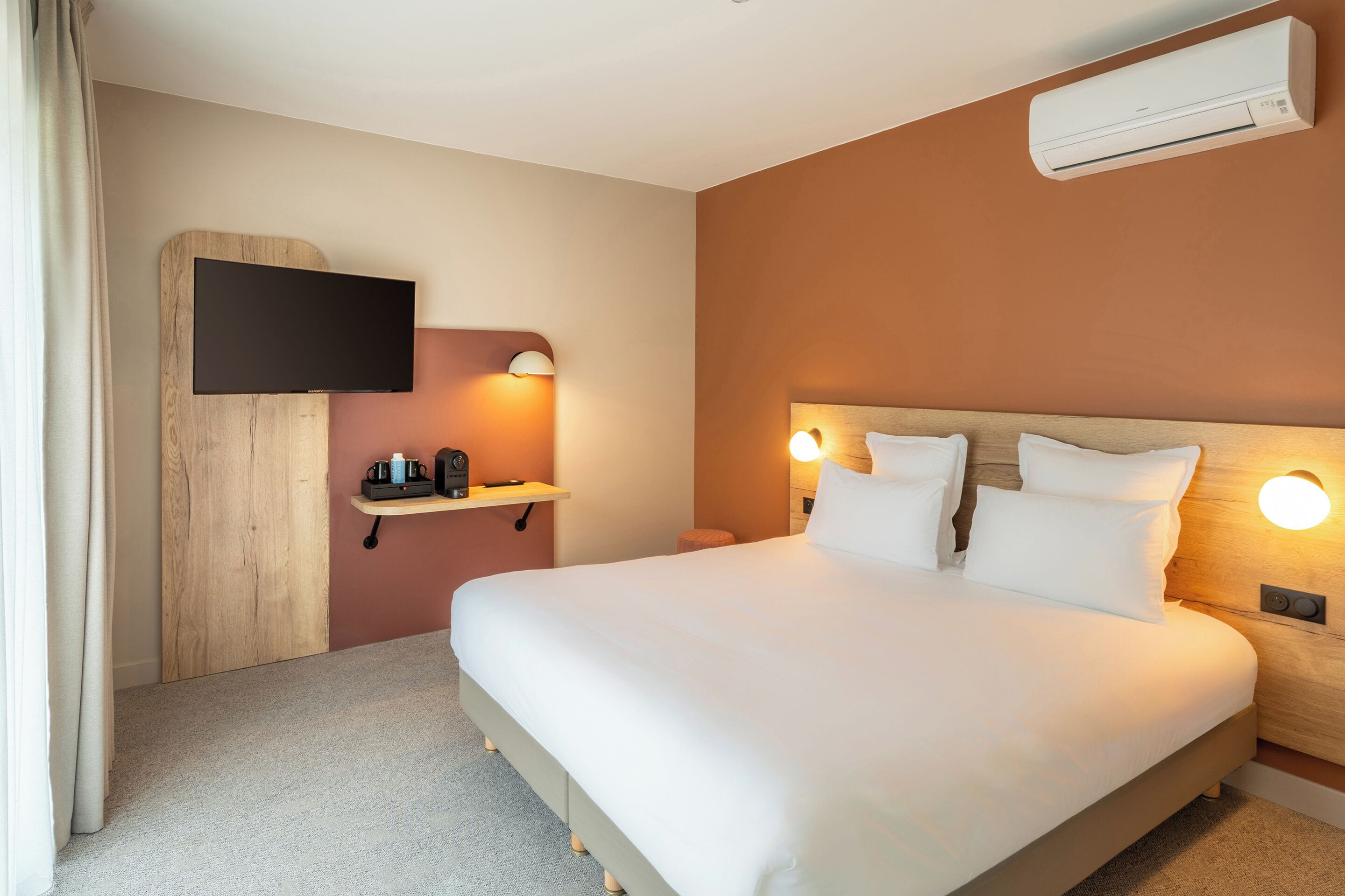 Habitación de hotel con una cama grande, un televisor de pantalla plana montado en un panel de madera, una mesita de noche con lámpara y un equipo de aire acondicionado.