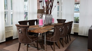 Een eetgedeelte met een houten tafel, zes gestoffeerde stoelen, een vaas met takken en roze kaarsen, en een raam met uitzicht op gebouwen.