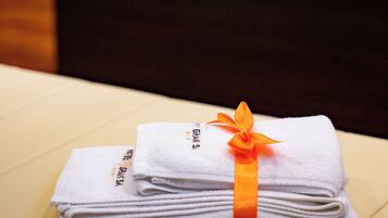 A neatly folded stack of white towels with an orange ribbon, placed on a beige surface in a room with a dark-colored sofa and a white cushion in the background.