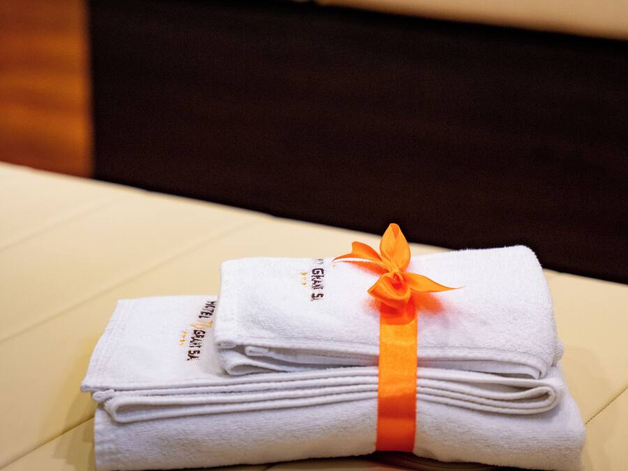 A neatly folded stack of white towels with an orange ribbon, placed on a beige surface in a room with a dark-colored sofa and a white cushion in the background.