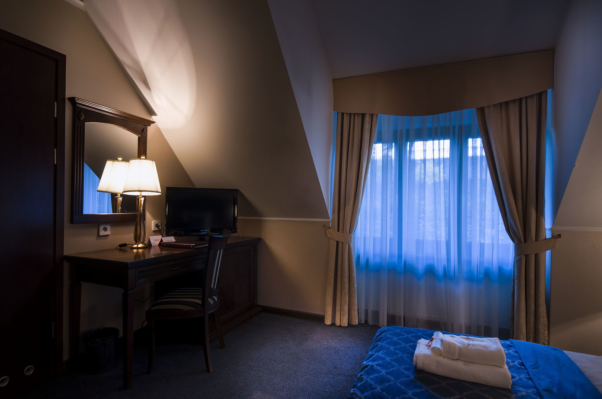 A hotel room with a desk, chair, television, and a bed with a blue bedspread and white towels.