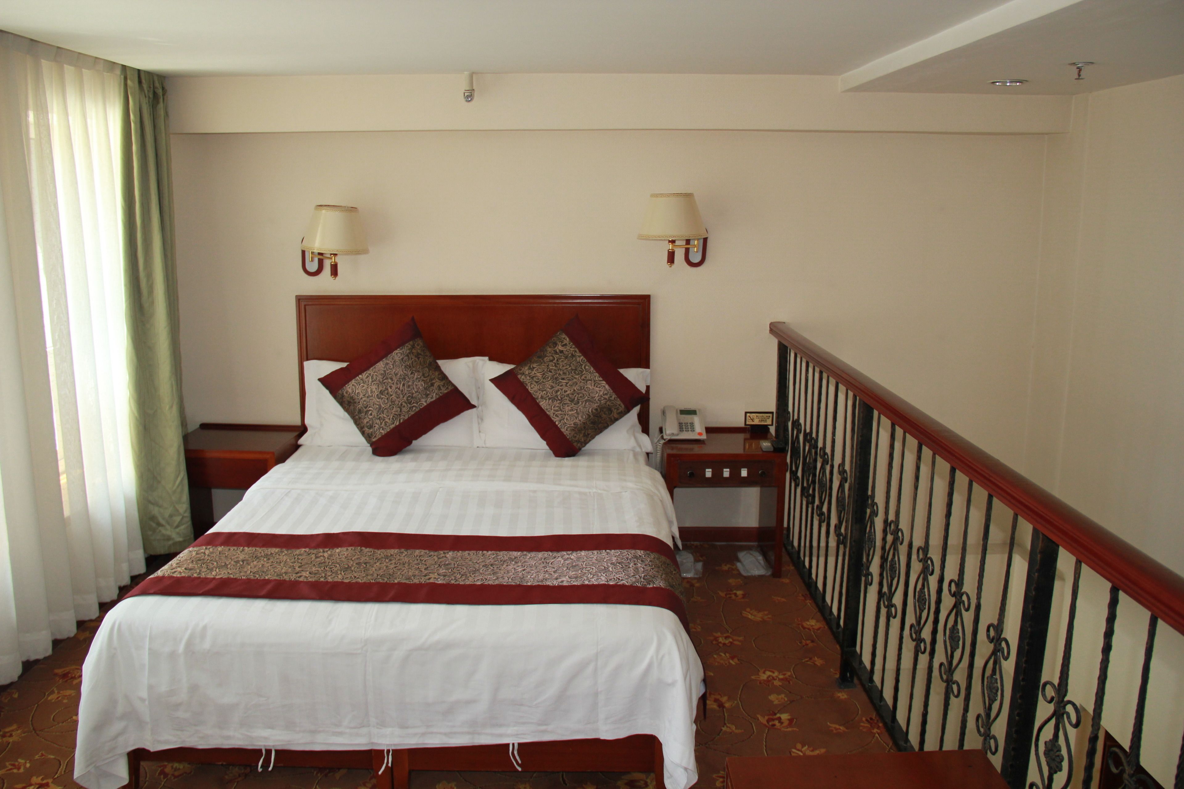 Une chambre d’hôtel avec un grand lit, deux tables de chevet, une petite table, une lampe et une rambarde le long de la fenêtre.