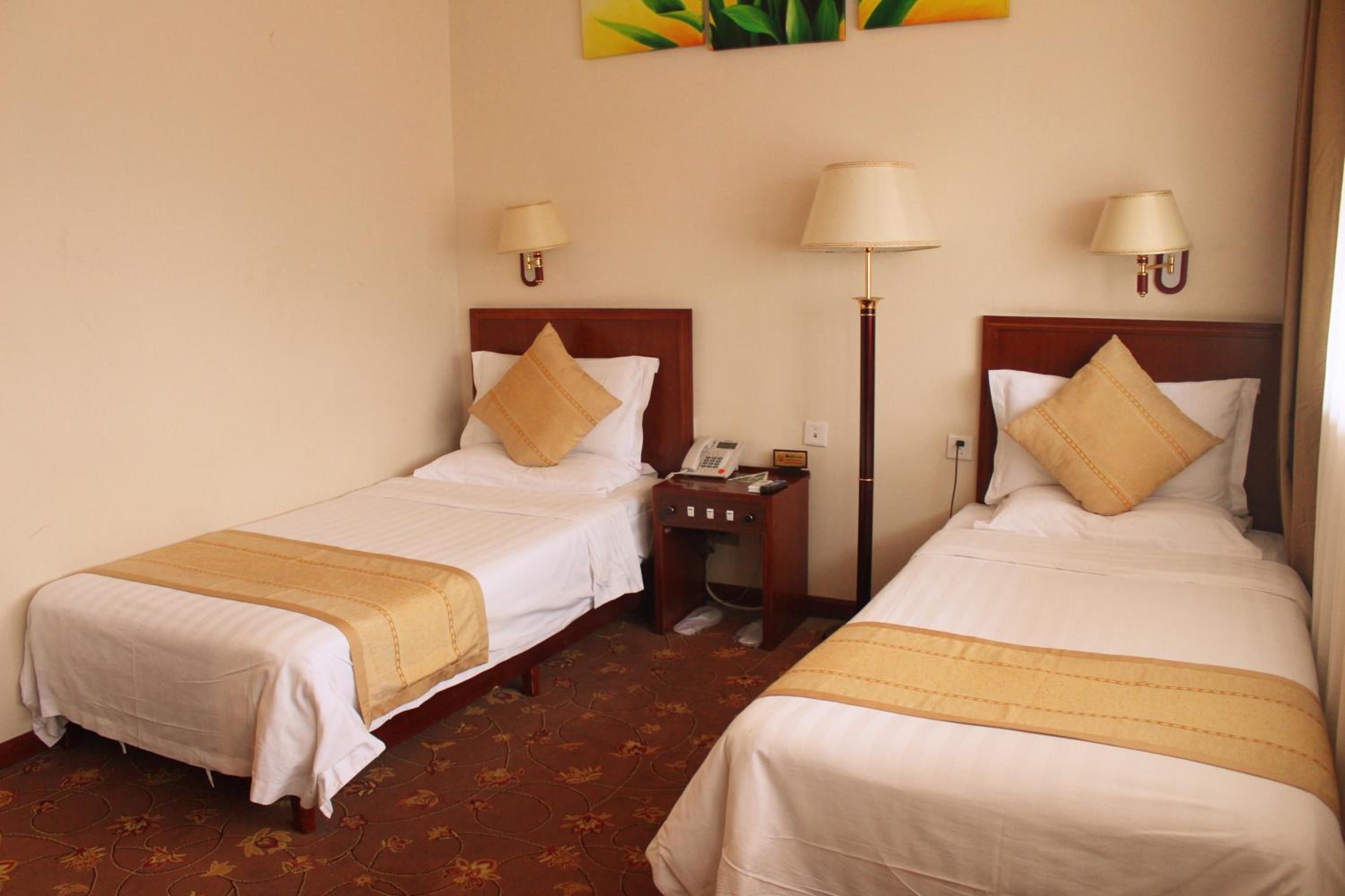 A hotel room with two single beds, each with a pillow and a bedside table with a lamp.
