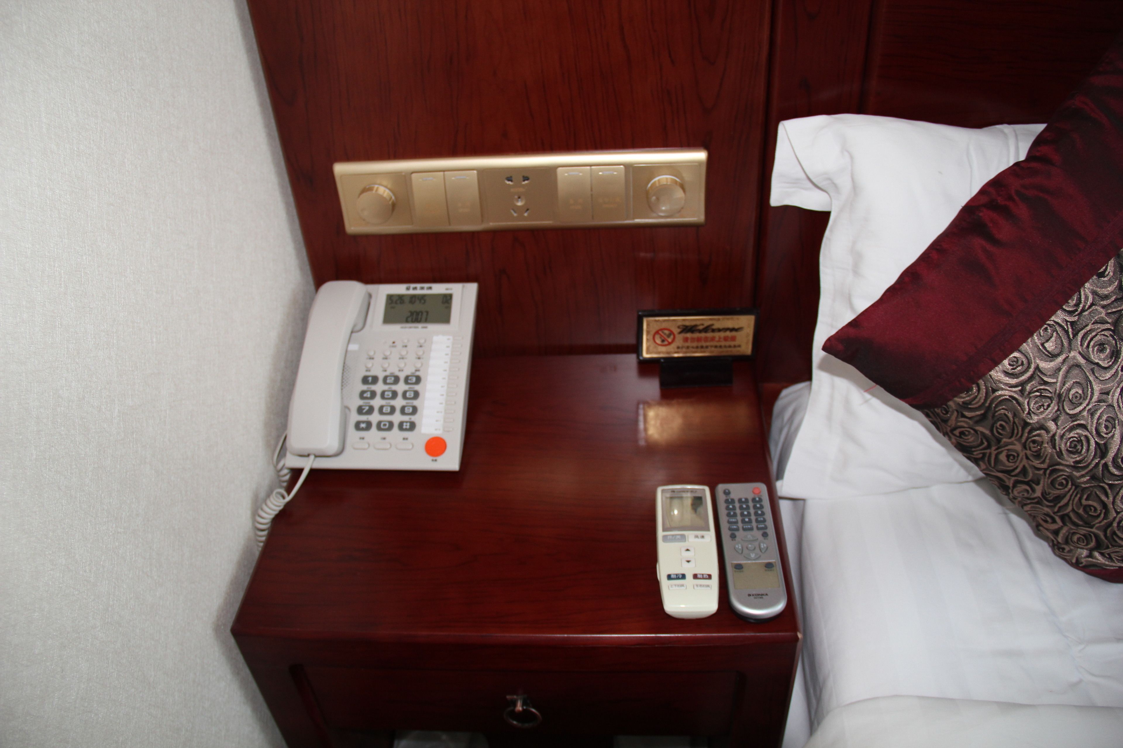A hotel room with a wooden nightstand, a white telephone, a digital clock, a remote control, and a bed with white and patterned pillows.