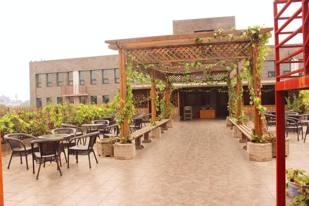 An outdoor patio with tables and chairs, a wooden pergola with climbing plants, and a building in the background.