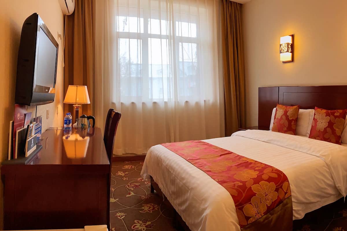 A hotel room with a bed, a desk with a lamp, a TV, and a window with curtains.