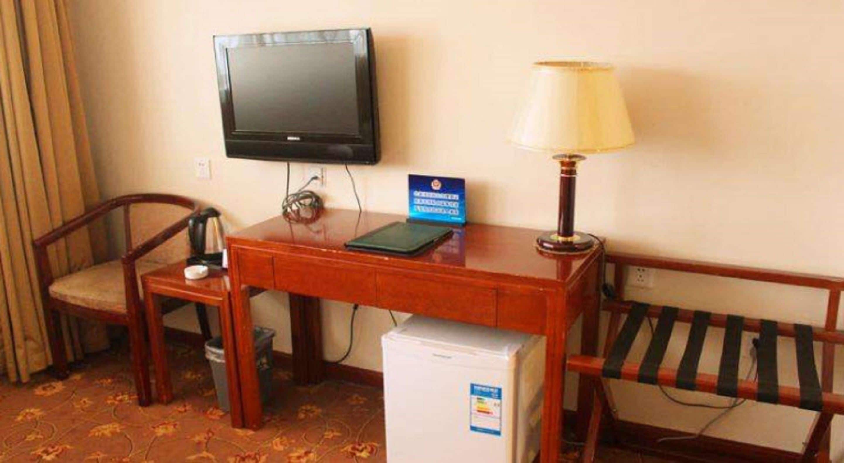 A hotel room with a wooden desk, a television, a lamp, a small refrigerator, a chair, and a bed with striped bedding.