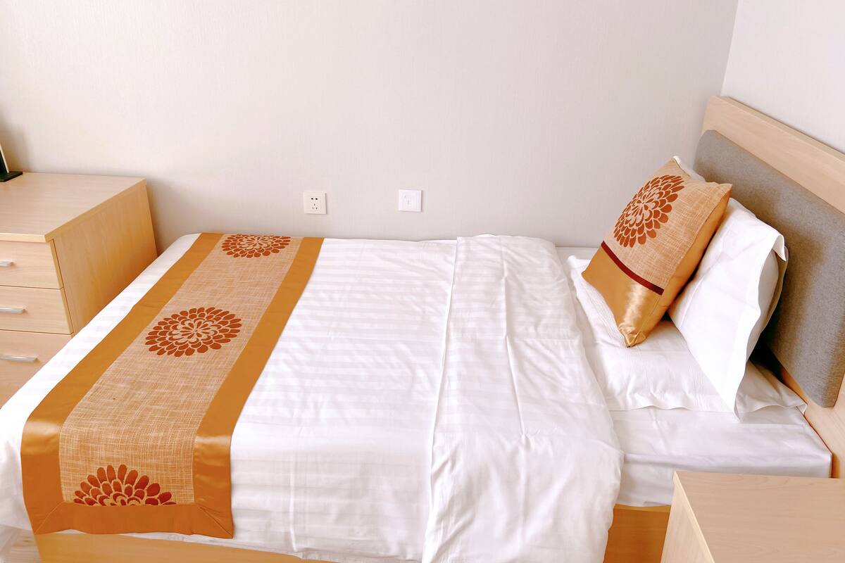 A neatly made bed with a white and orange bedspread, a wooden nightstand, and a plain wall with two electrical outlets.