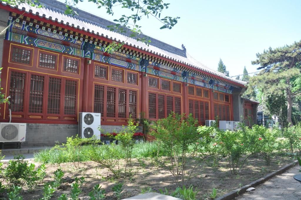A traditional building with a tiled roof and red walls, surrounded by a garden with small trees and shrubs.