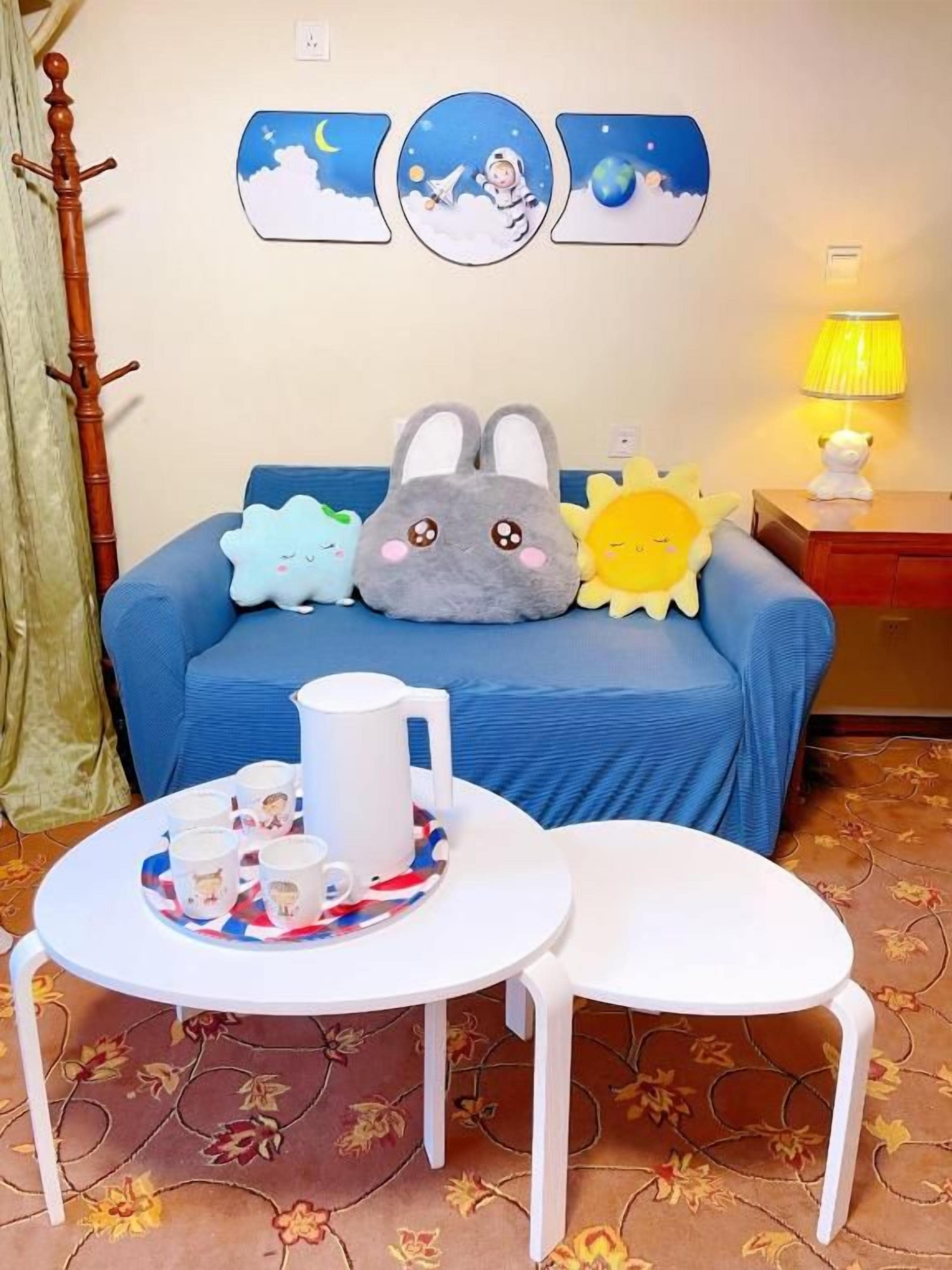 A room with a blue sofa, two pillows, a small table with a pitcher and cups, and a lamp.