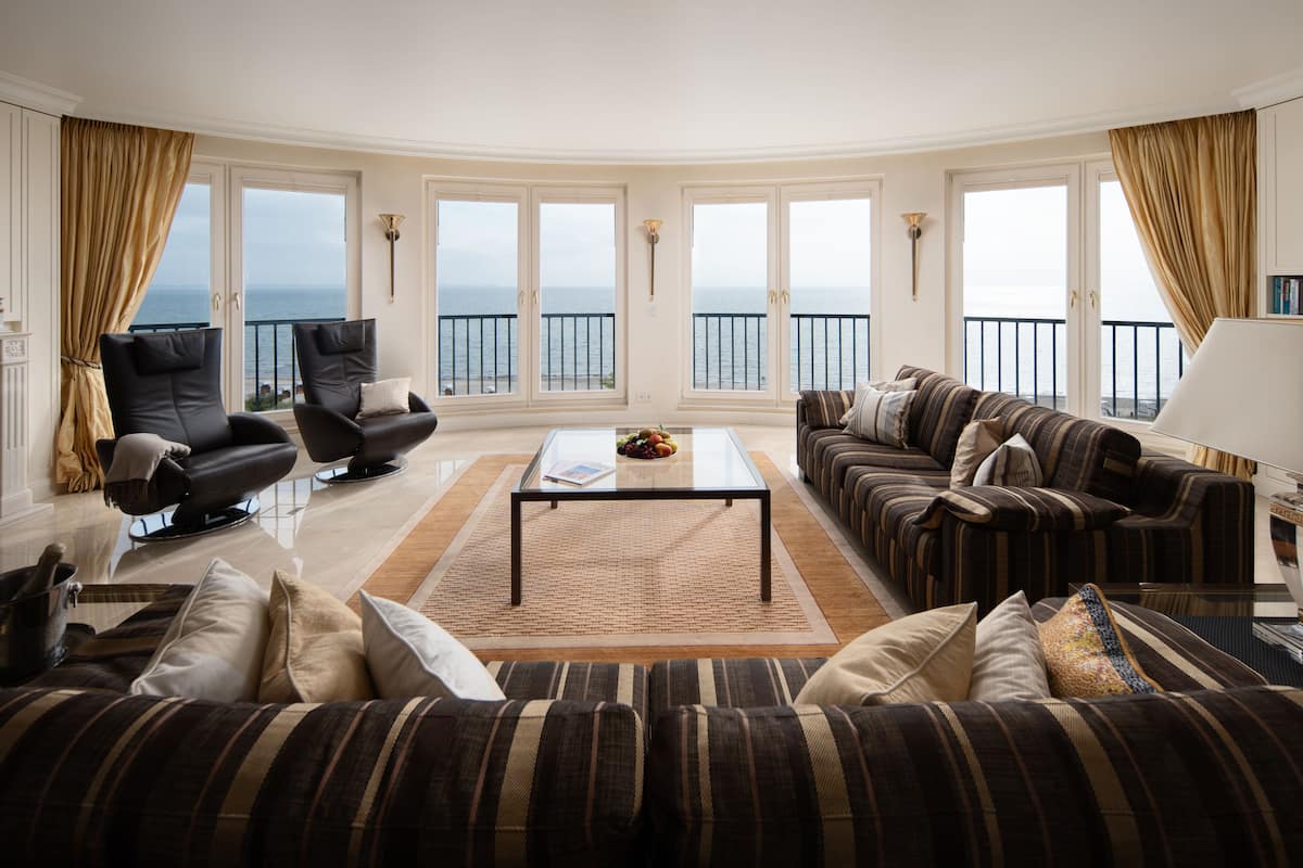 Uma sala de estar espaçosa com uma grande janela com vista para o mar, um sofá, uma mesa de centro e duas poltronas.