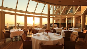 Un restaurante con mesas redondas dispuestas para comer, amplias ventanas que ofrecen vistas de la puesta de sol, y un diseño de interiores moderno.