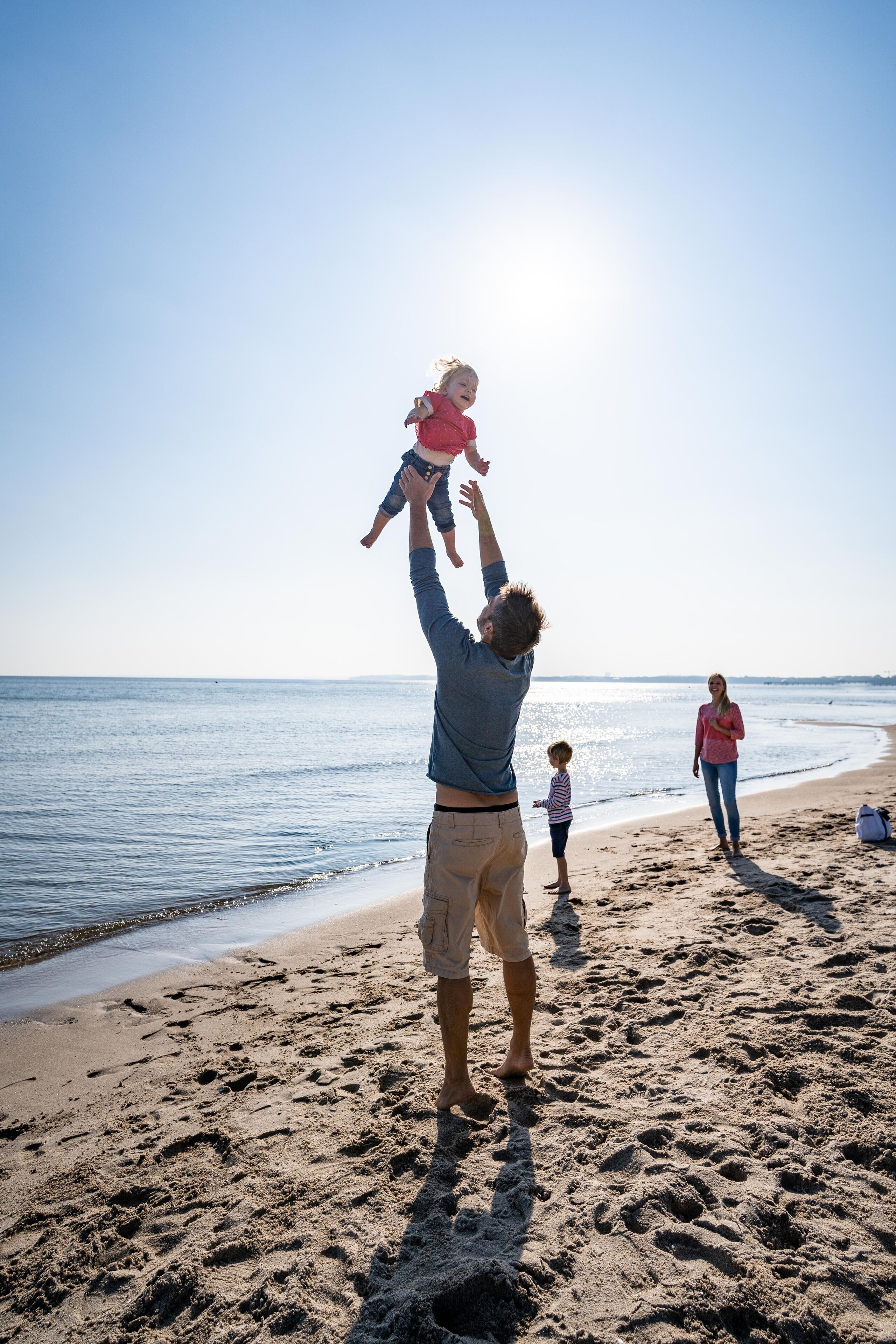 Escena de playa con una persona levantando a un niño, otro adulto de pie al fondo y un niño caminando.