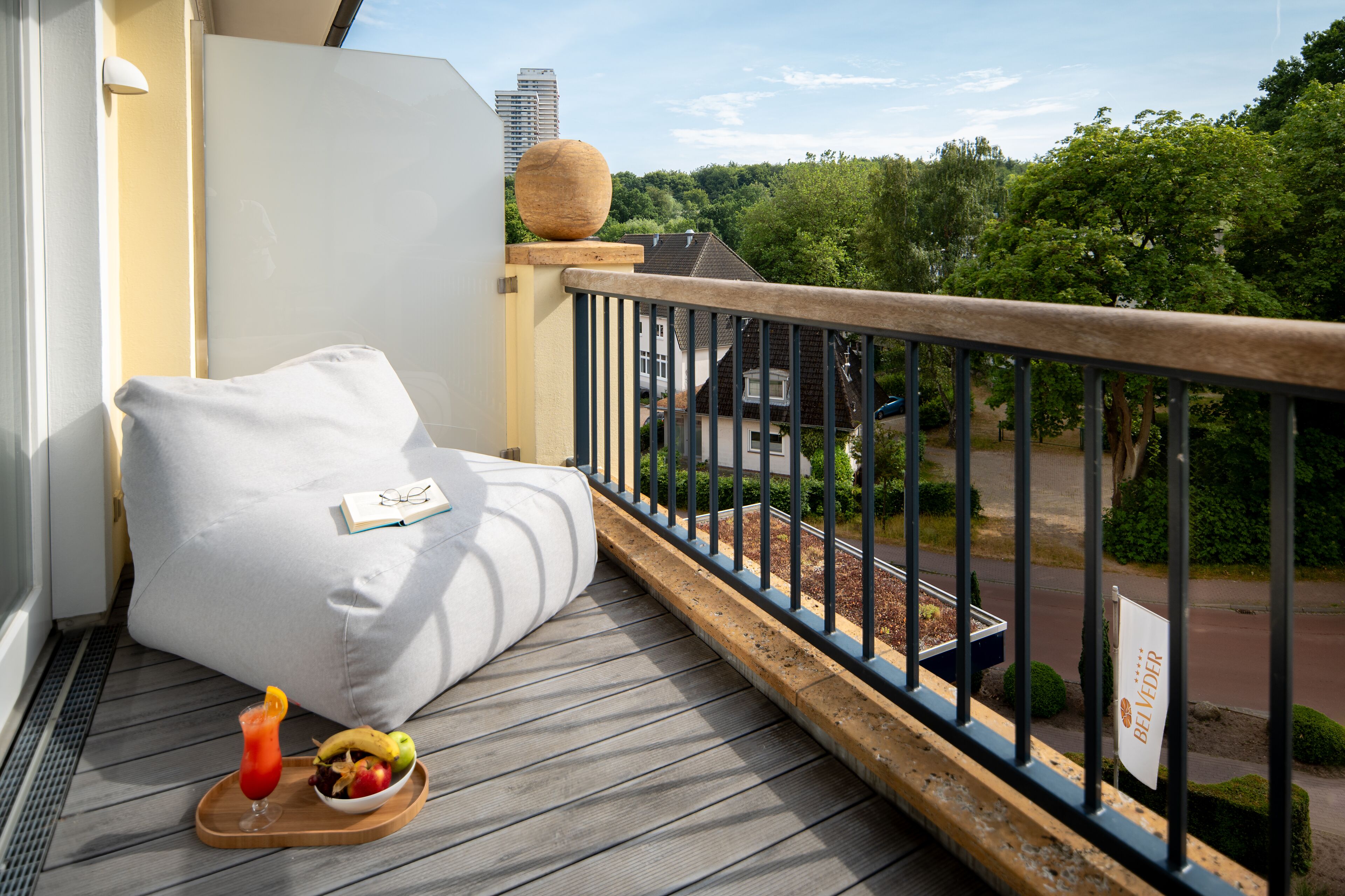 Ein Balkon mit weißem Kissen, einem Tablett mit Getränken und Früchten sowie Blick auf eine Straße und Grünflächen.