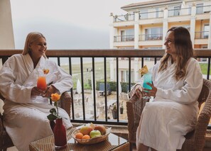 Dos personas con batas blancas sentadas en sillas de mimbre en un balcón con vista a un edificio y un jardín.