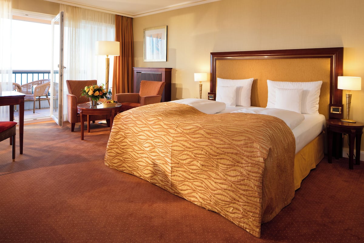 Quarto de hotel com uma cama grande, área de estar com uma poltrona e mesa, e vista para o exterior.