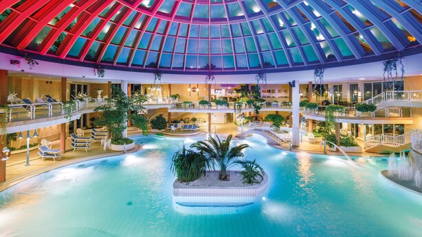 Una piscina interior de varios niveles con techo abovedado de vidrio, múltiples zonas de descanso y una isla central con plantas.