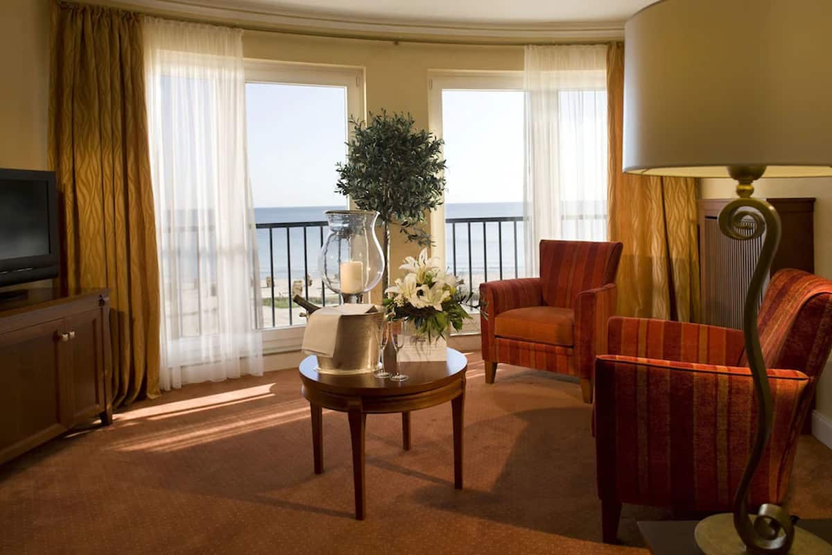 Quarto de hotel com varanda, televisão, mesa com um vaso de flores e poltrona listrada.