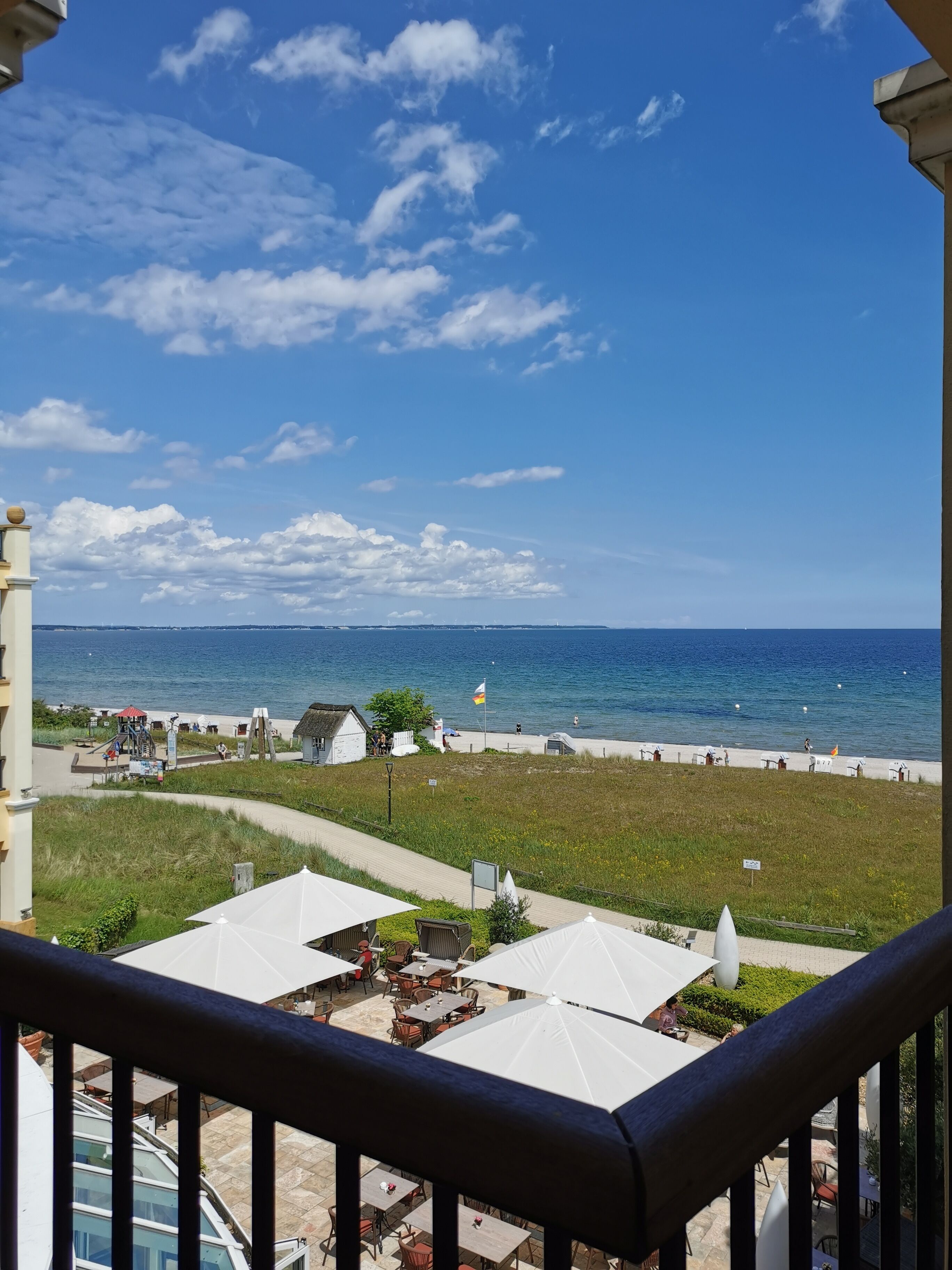 A seaside view from a balcony with outdoor seating, a beach, and a clear sky.
