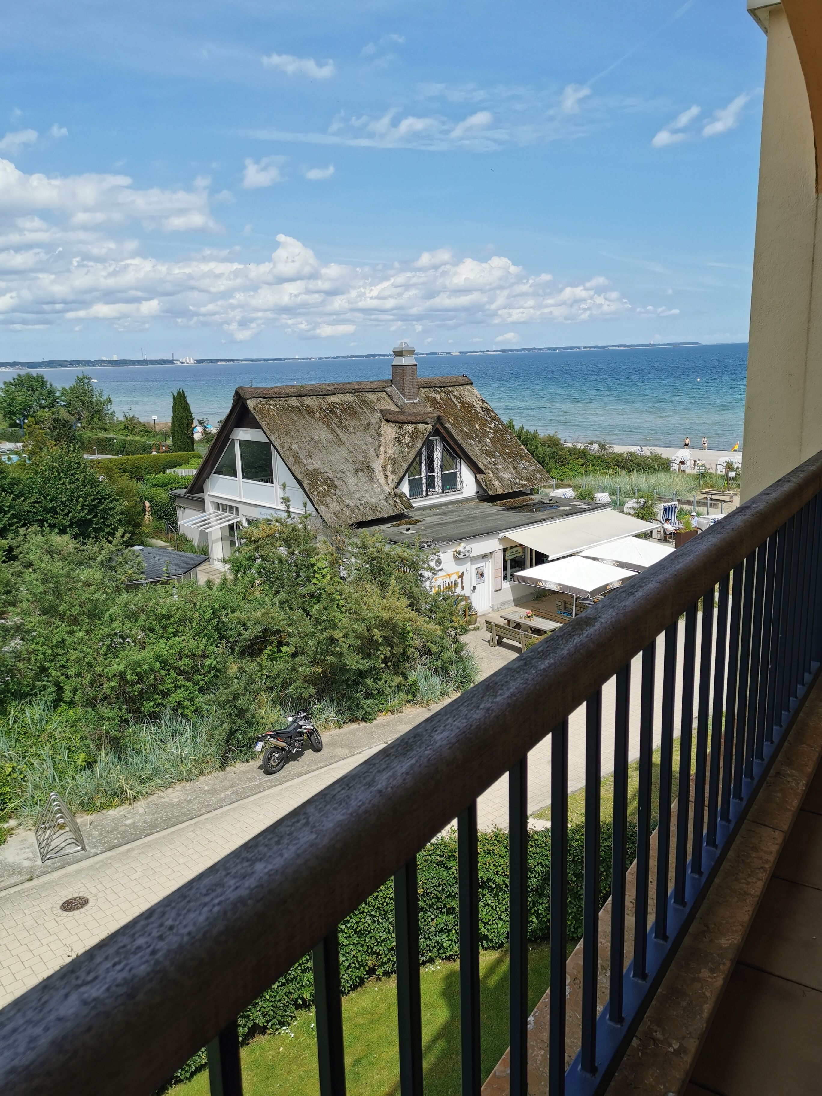 A coastal house with a thatched roof, a balcony with a wooden railing, and a view of the sea and distant buildings.