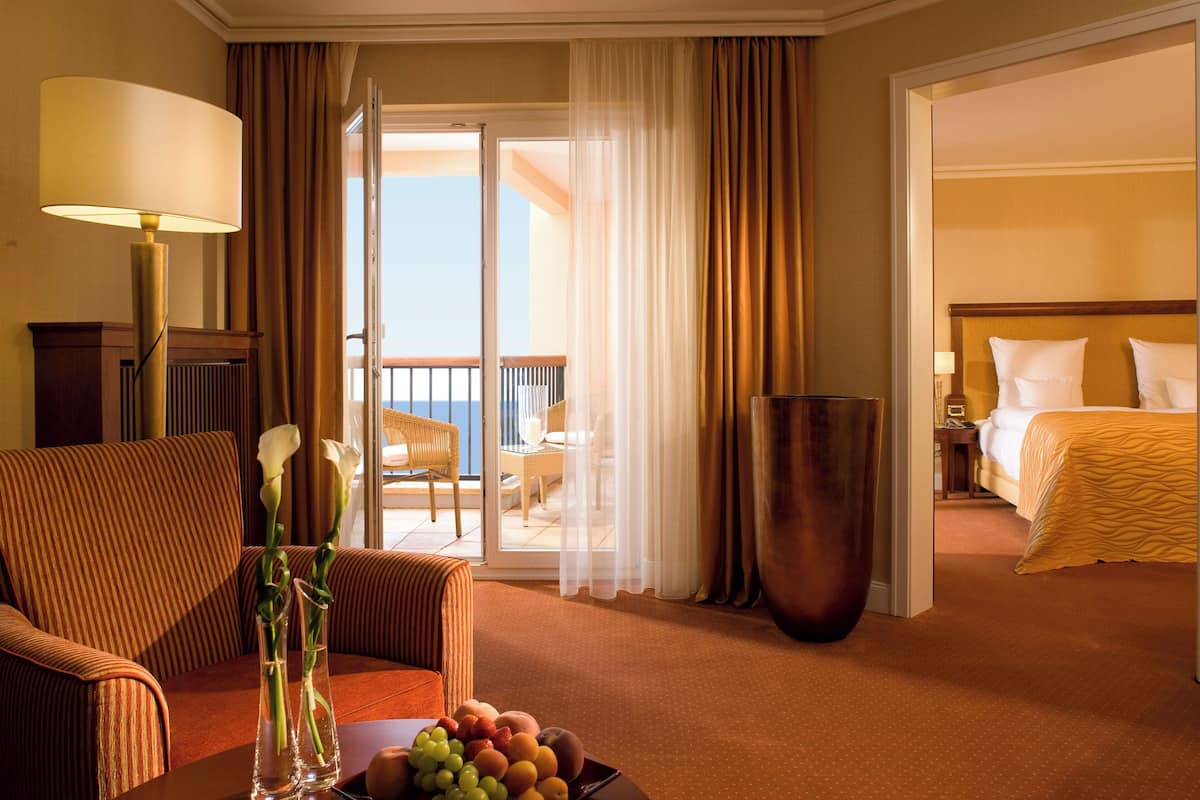 Quarto de hotel com uma cama, área de estar com uma cadeira e uma mesa, um vaso grande com flores e vista para a varanda externa.
