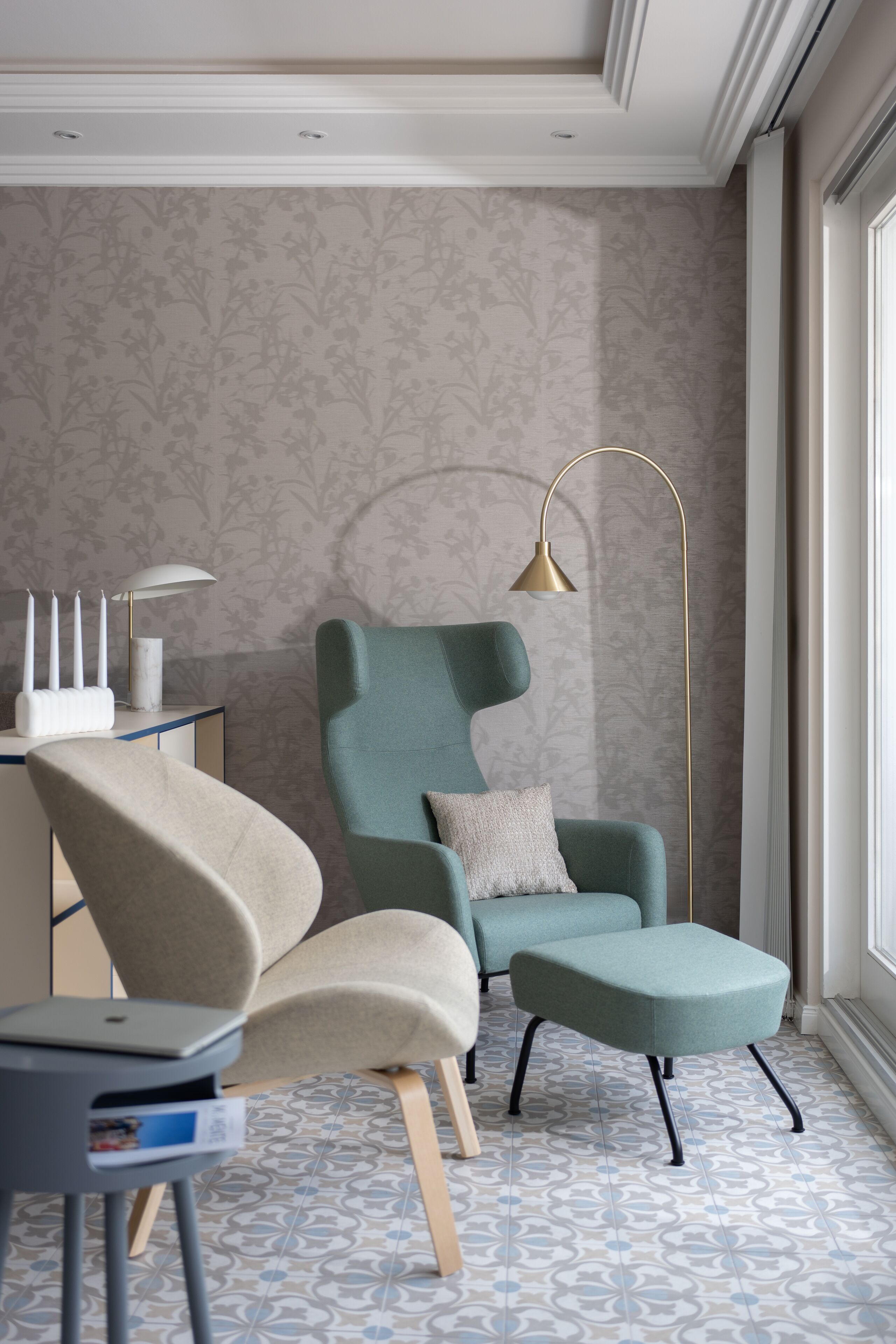 En moderne stue med en teal farvet lænestol, et matchende fodskammel, et sofabord med en lampe og tapet med mønster.