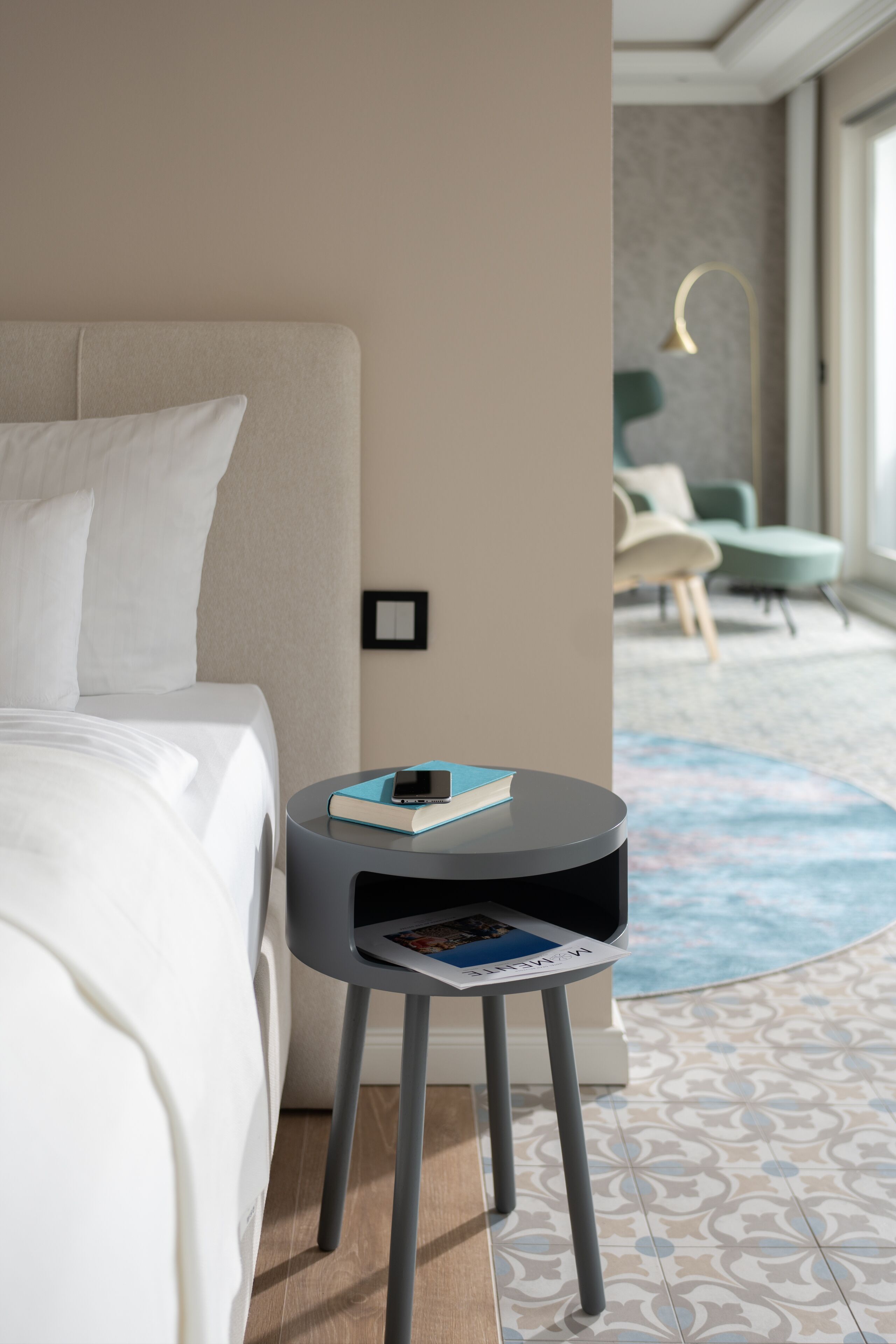 A modern hotel room with a neatly made bed, a small side table with a book and a magazine, and a view of the room's interior.