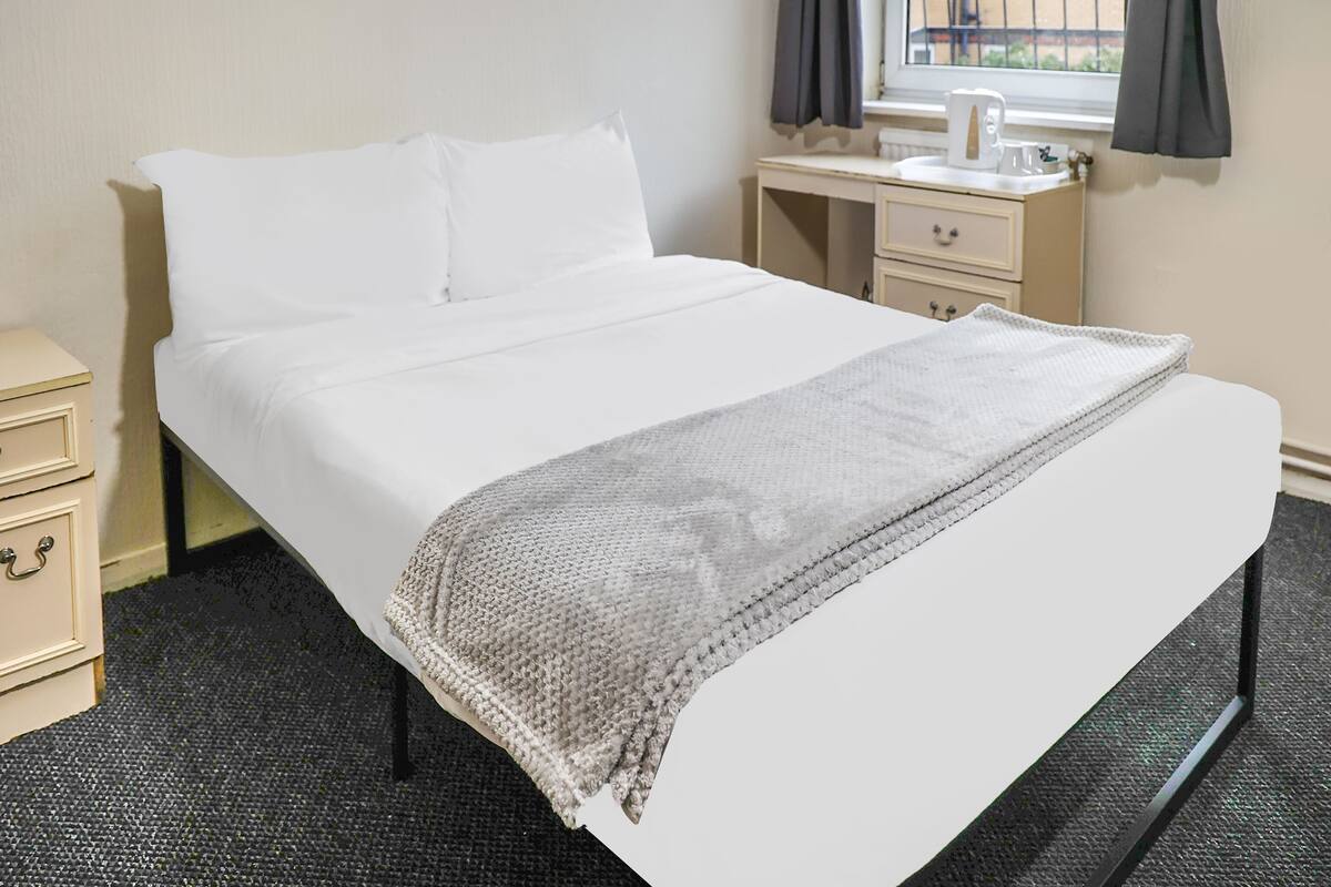 A neatly made bed with white bedding and a gray knitted throw, a small bedside table with a lamp, and a window with curtains.