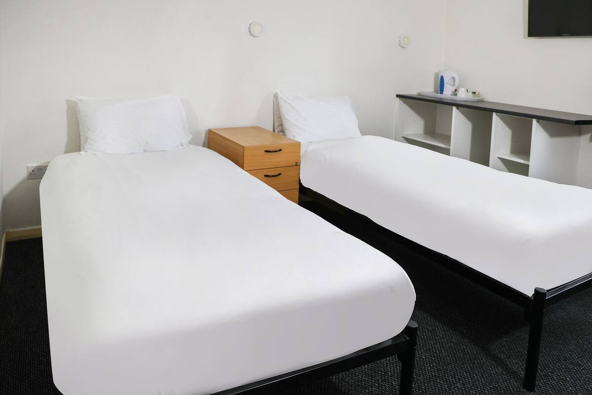 A hotel room with two single beds, a wooden dresser, a small table with a kettle, and a shelf unit.