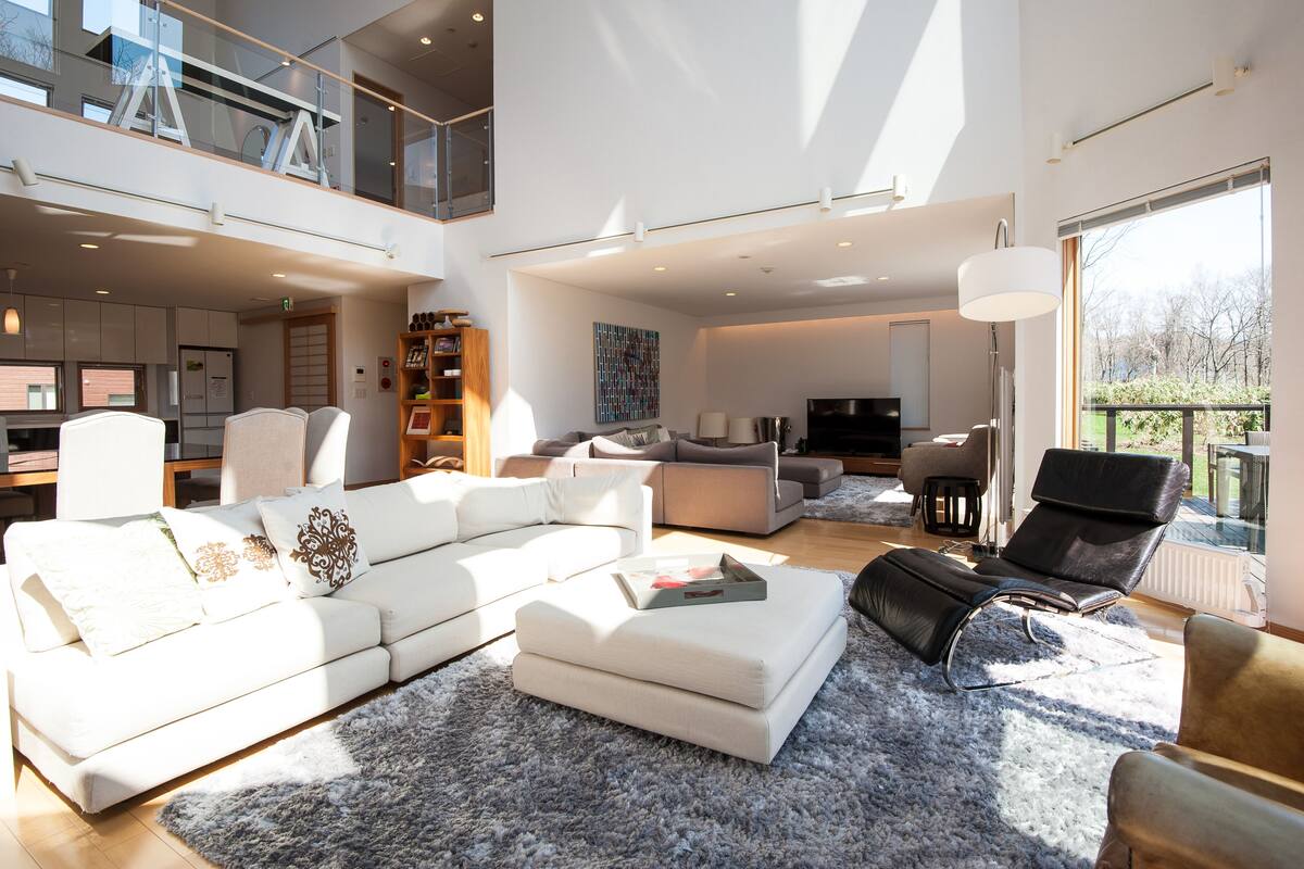 A spacious living area with a white sectional sofa, a black leather chair, and a large window offering a view of the outdoors.
