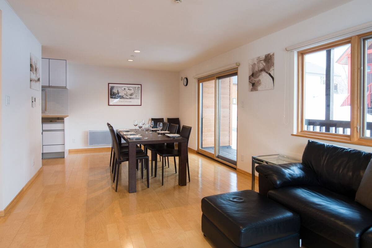 A modern living room with a black leather sofa, a dining table with chairs, and a kitchen area with cabinets and a window.