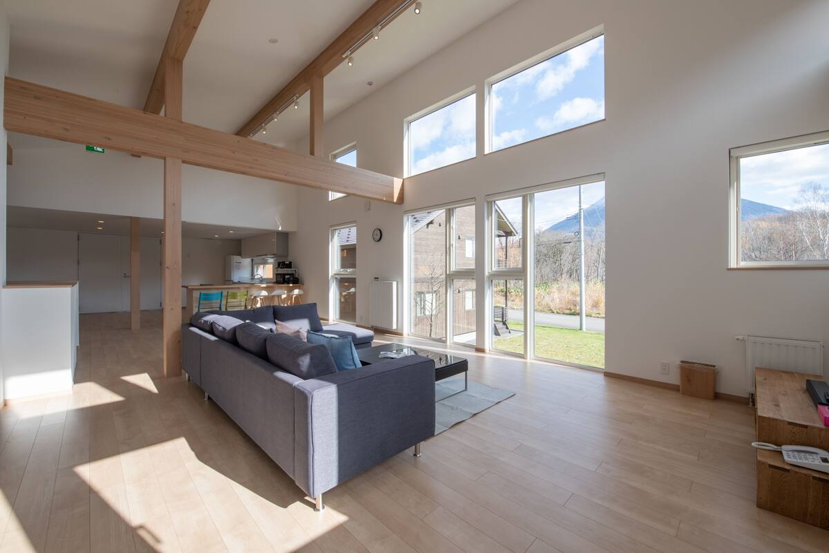 A spacious living area with a large gray sofa, wooden beams, and large windows offering a view of the outdoors.