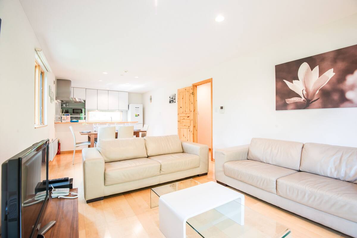 A modern living room with two beige sofas, a glass coffee table, and a flat-screen TV. A kitchen with white cabinets and a dining area with a table and chairs is visible in the background.